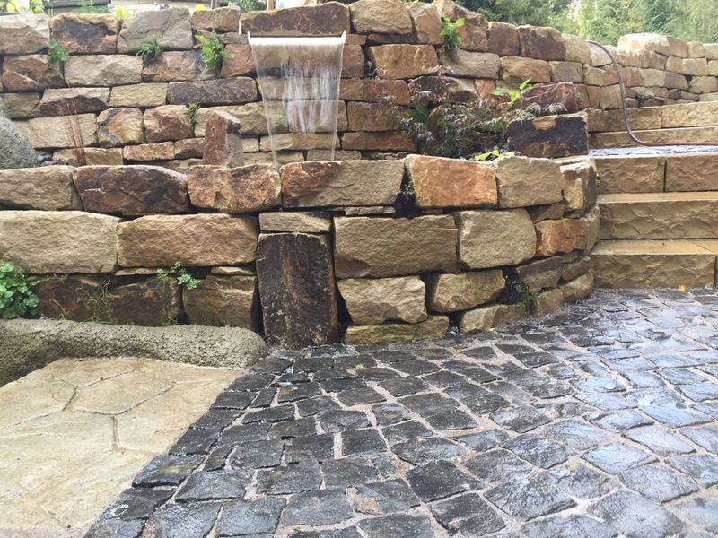 Ein gemauerter Wasserfall aus Natursteinen, der in ein Kopfsteinpflasterbecken mündet.